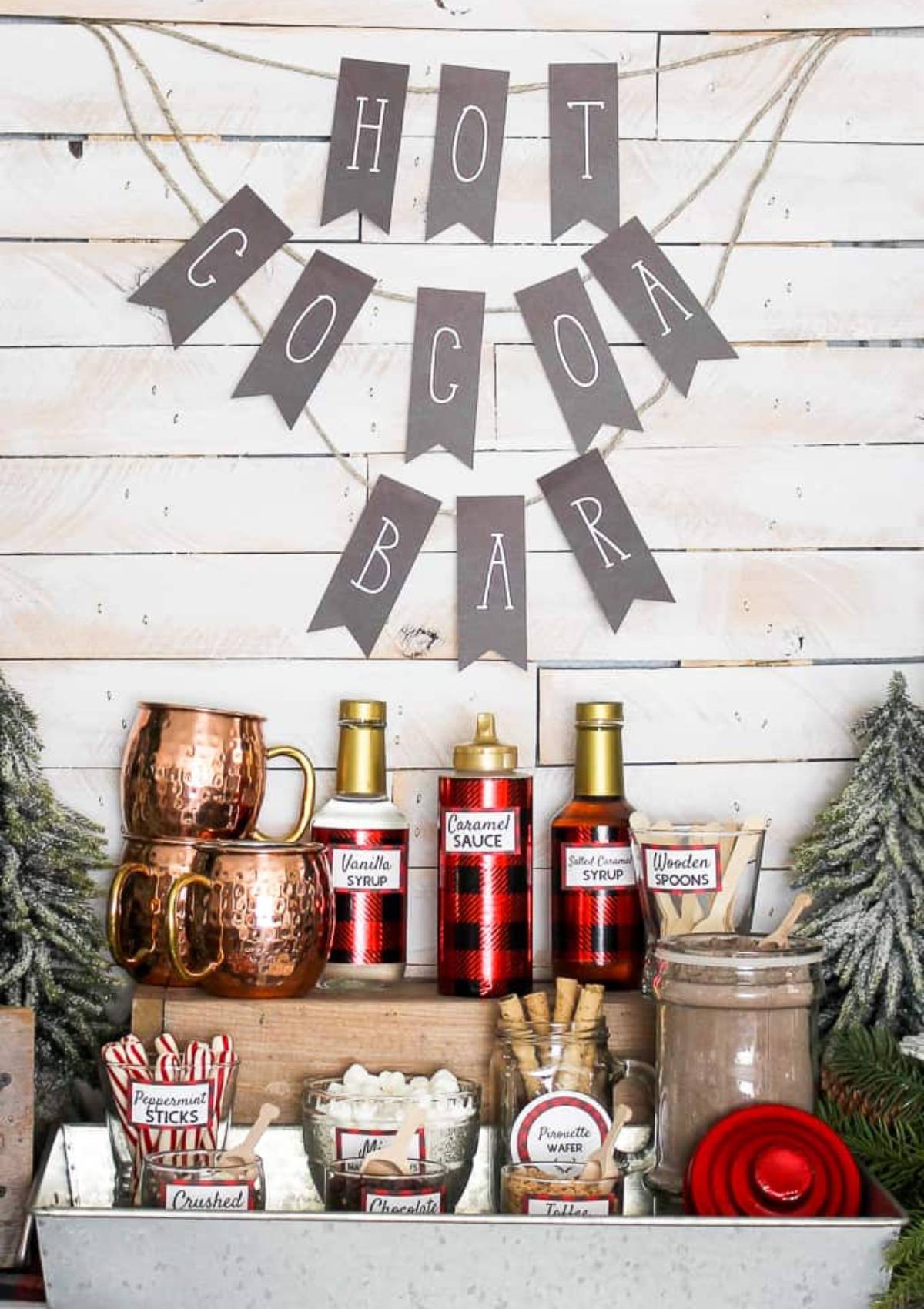 A festive hot cocoa bar with a banner that says "Hot Cocoa Bar." The bar includes mugs, syrups, marshmallows, candy canes, and other toppings.