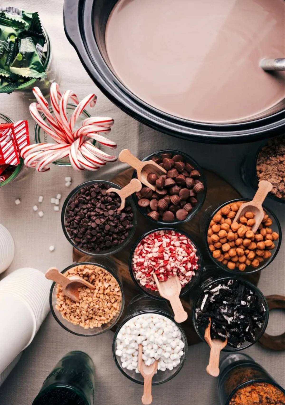 A festive hot chocolate bar with a slow cooker filled with hot chocolate in the center. Surrounding the slow cooker are various toppings in small bowls, including marshmallows, chocolate chips, candy canes, sprinkles, and nuts. There are also small scoops and paper cups on the table.