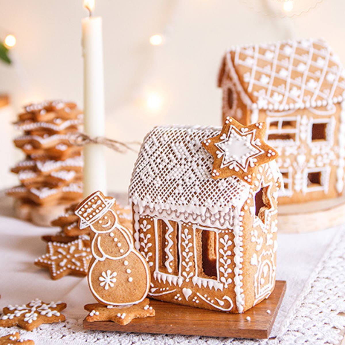 A charming gingerbread house adorned with intricate icing details stands on a wooden board. A gingerbread snowman stands proudly beside it, and several other festive cookies, including stars, add to the scene.