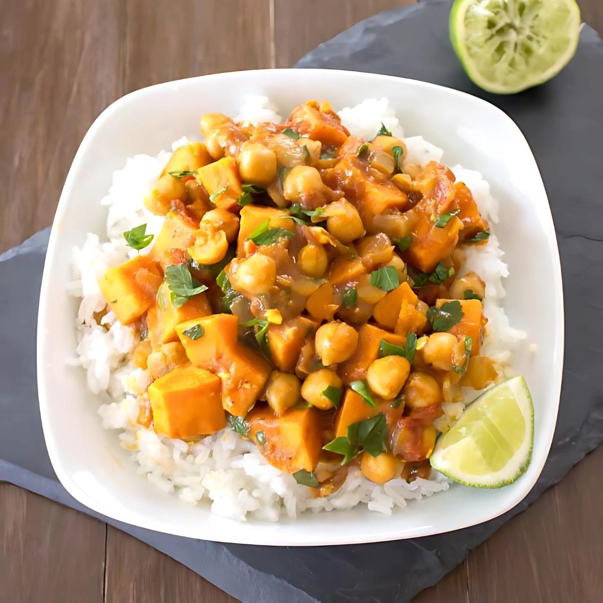 A plate of curry with chickpeas, sweet potatoes, and rice, garnished with fresh parsley and lime wedges.