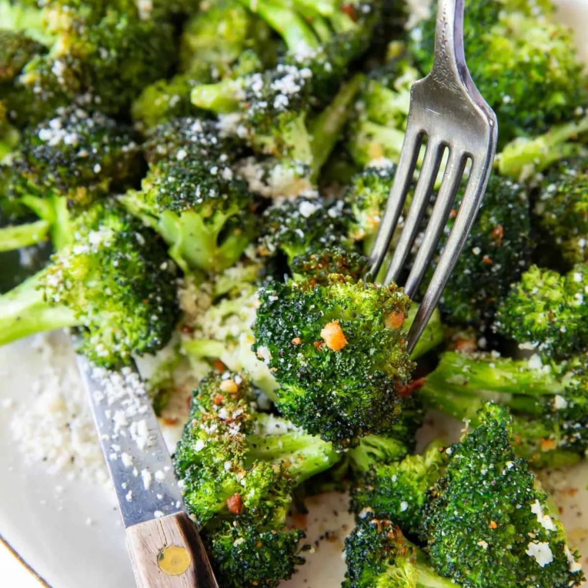 A close-up of a plate of air fried broccoli with a fork scooping up a piece. The broccoli florets are bright green and look crispy and seasoned with grated cheese and red pepper flakes.