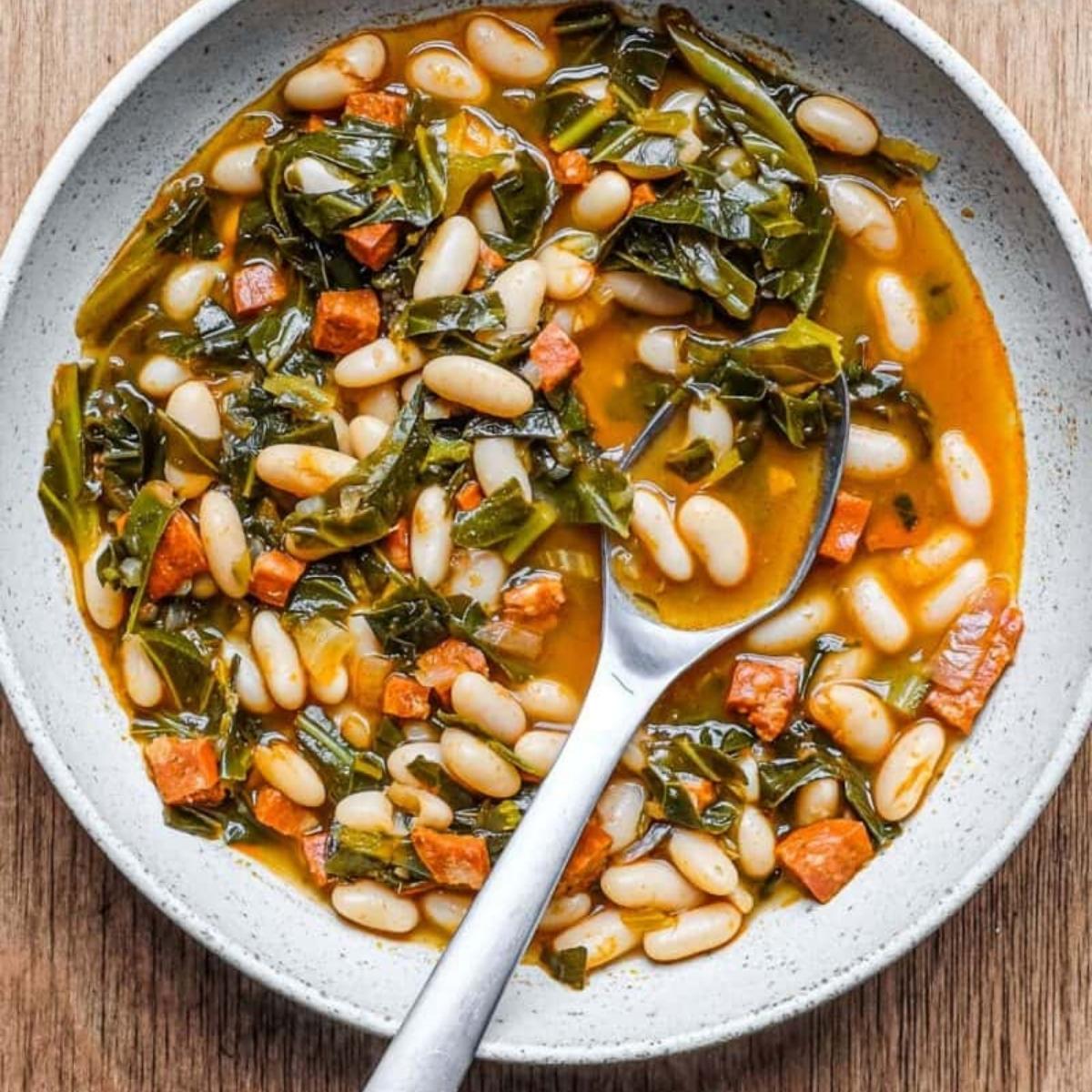 A bowl of hearty, rustic soup with white beans, kale, and chorizo. The soup looks flavorful and comforting, with a rich broth and a generous amount of vegetables and sausage.