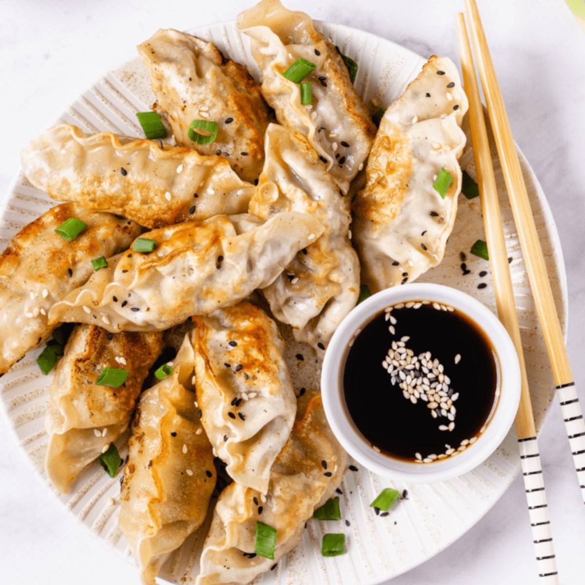 A plate of golden-brown dumplings with a side of dipping sauce and chopsticks. The dumplings are sprinkled with sesame seeds and green onions.