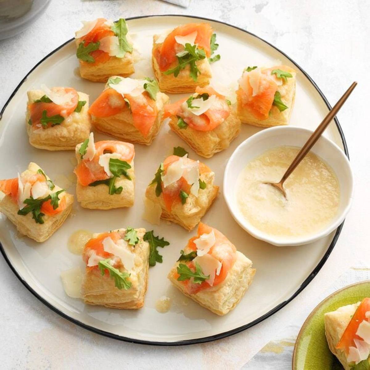 A plate of savory appetizers featuring flaky puff pastry squares topped with smoked salmon, arugula, tomato, and Parmesan cheese. A small bowl of creamy sauce is also included.