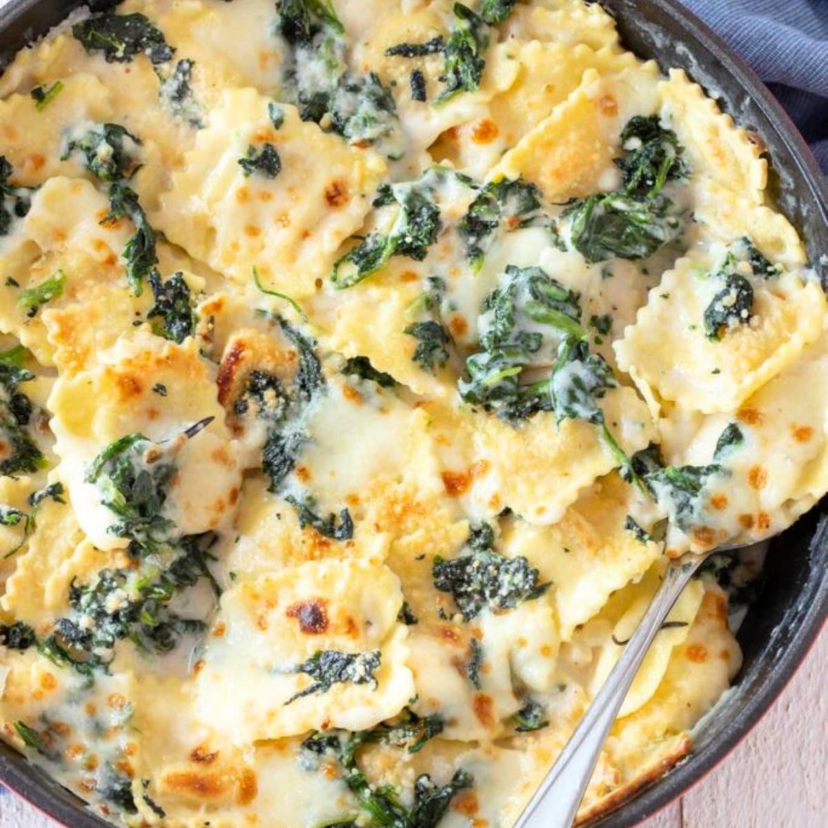 A skillet filled with a bubbly, cheesy spinach and ravioli bake. The ravioli is coated in a creamy sauce and topped with melted cheese, creating a delicious and comforting dish.