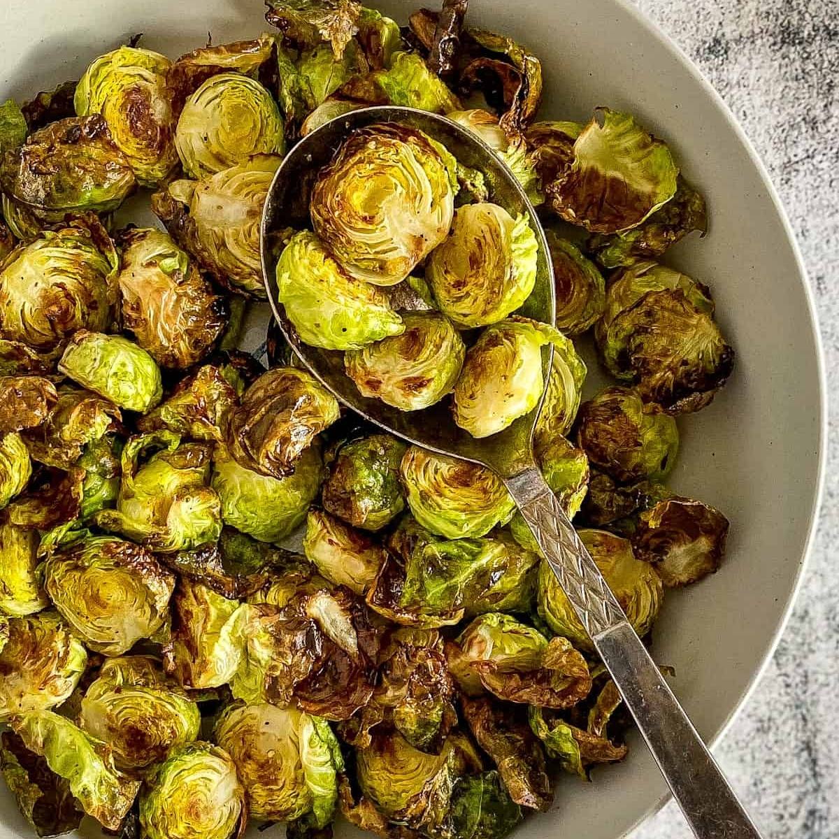 A bowl of air fried Brussels sprouts. The sprouts are golden brown and look crispy and delicious. A spoon is resting on top of the sprouts.