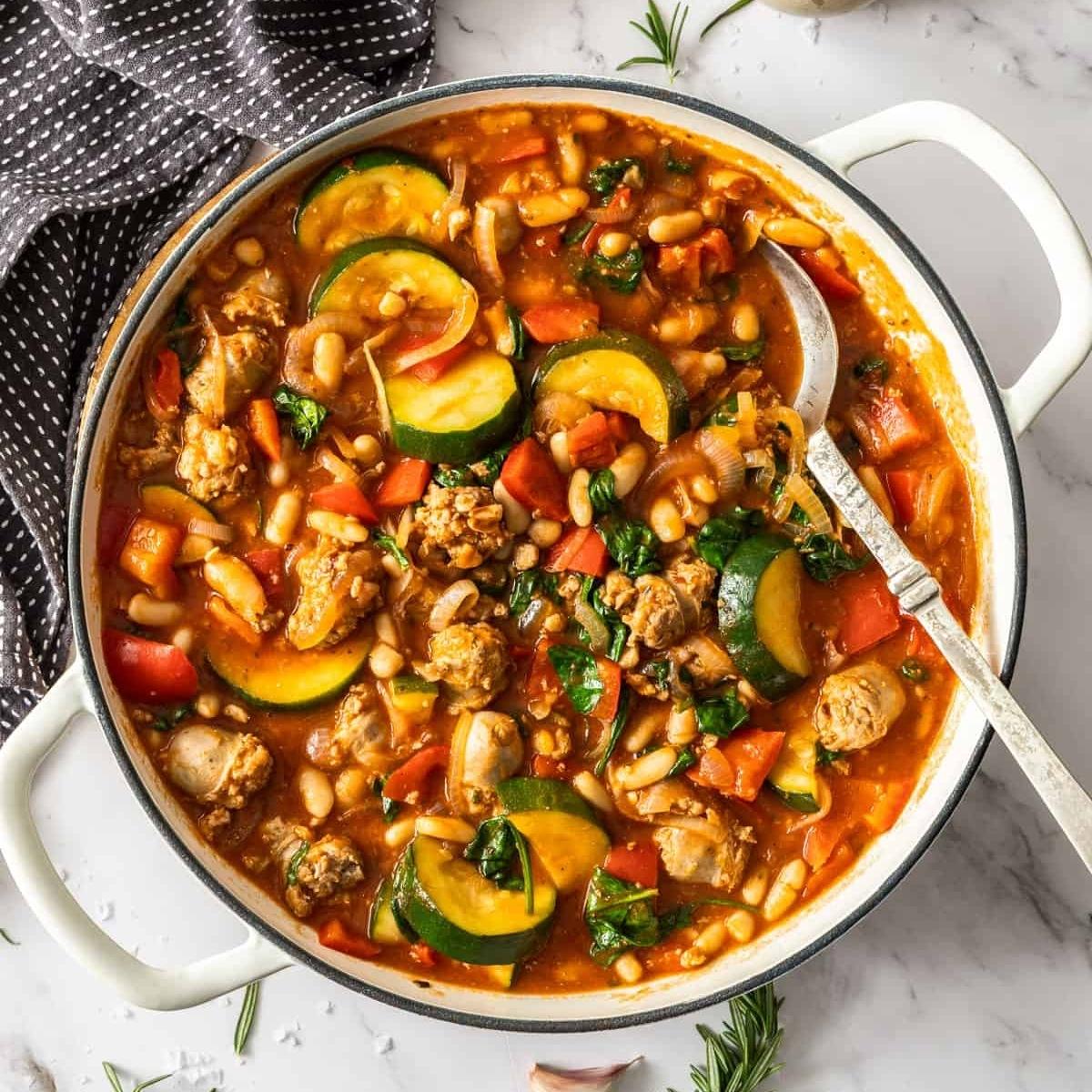 A hearty and colorful sausage and white bean stew simmering in a white pot. The stew is filled with vegetables like zucchini, peppers, and onions, and garnished with fresh parsley.