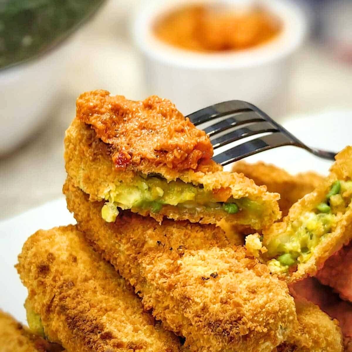 A fork lifting a crispy, golden-brown vegetable patty. The patty is filled with green peas and vegetables, and it's being dipped into a bowl of red sauce.