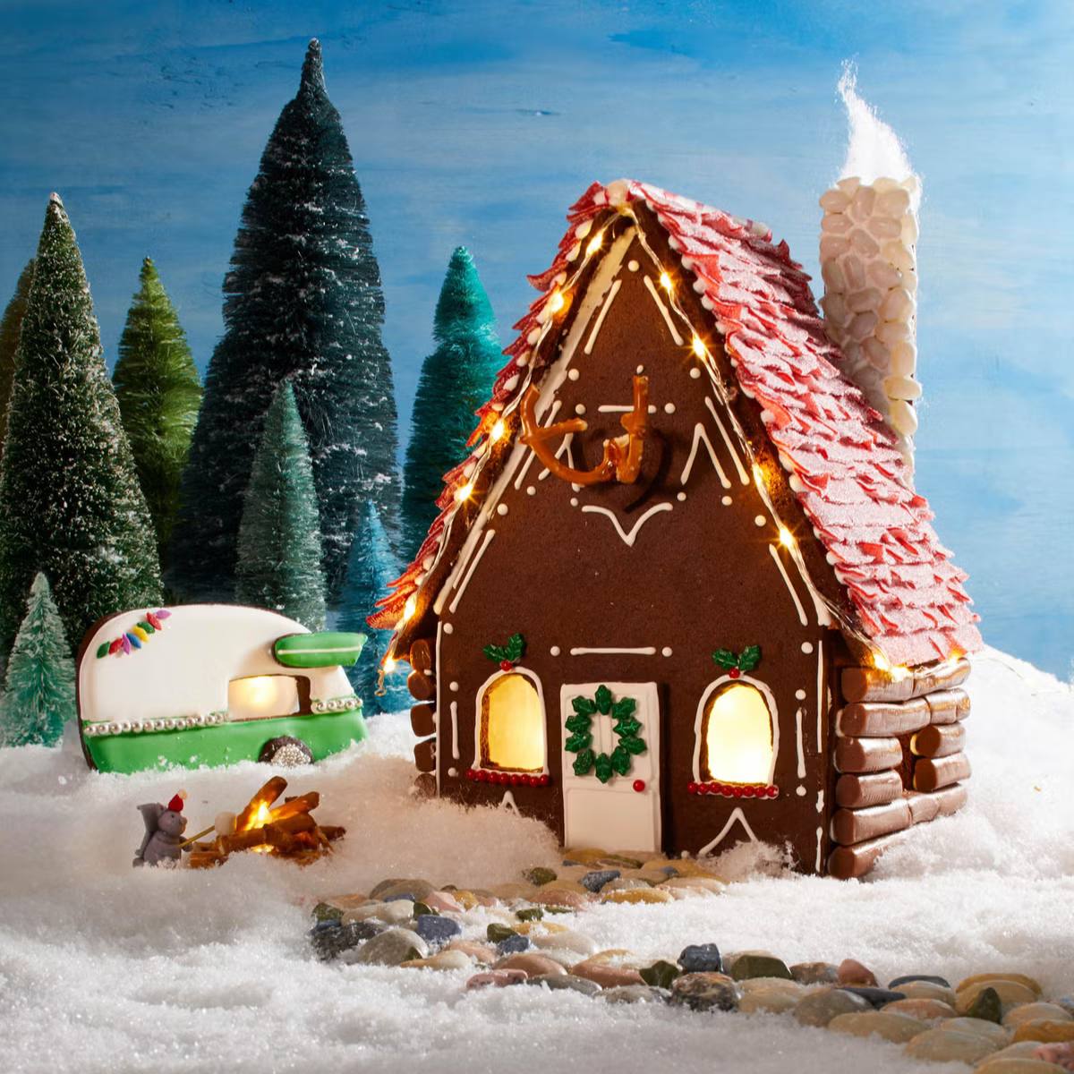 A whimsical gingerbread cabin with a lit chimney, a camper, and a campfire scene, all set against a snowy backdrop.