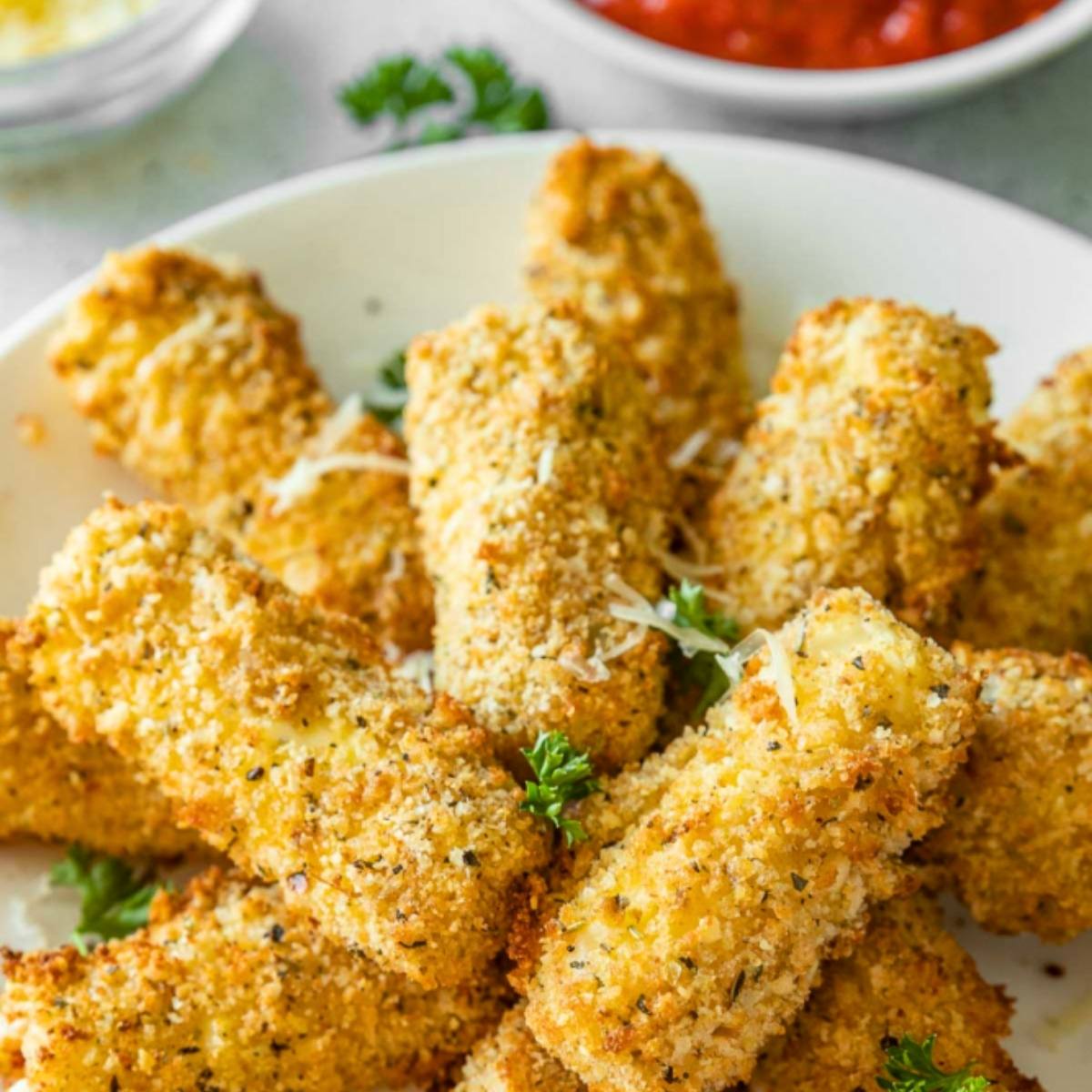 A plate of golden-brown, breaded mozzarella sticks. The sticks are sprinkled with grated cheese and parsley, and there are bowls of marinara sauce and grated cheese in the background.