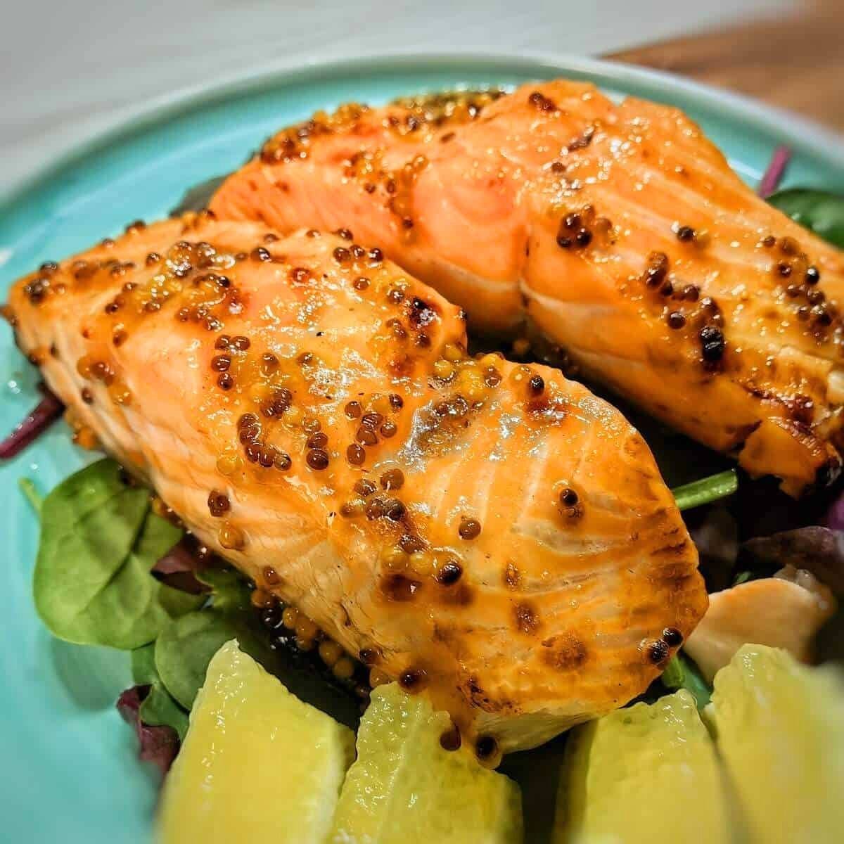 Two perfectly cooked salmon fillets on a plate. The salmon is glazed with a mustard sauce and is resting on a bed of greens and lemon slices.







