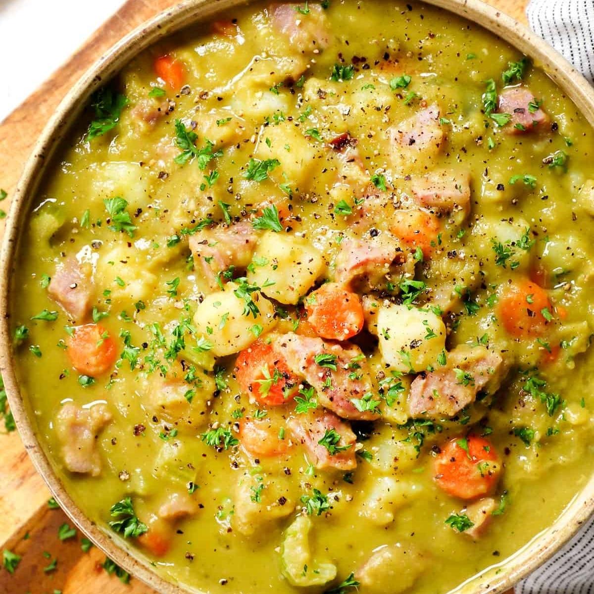 A bowl of creamy split pea soup with chunks of ham, carrots, and potatoes. The soup is garnished with fresh parsley and a sprinkle of black pepper.