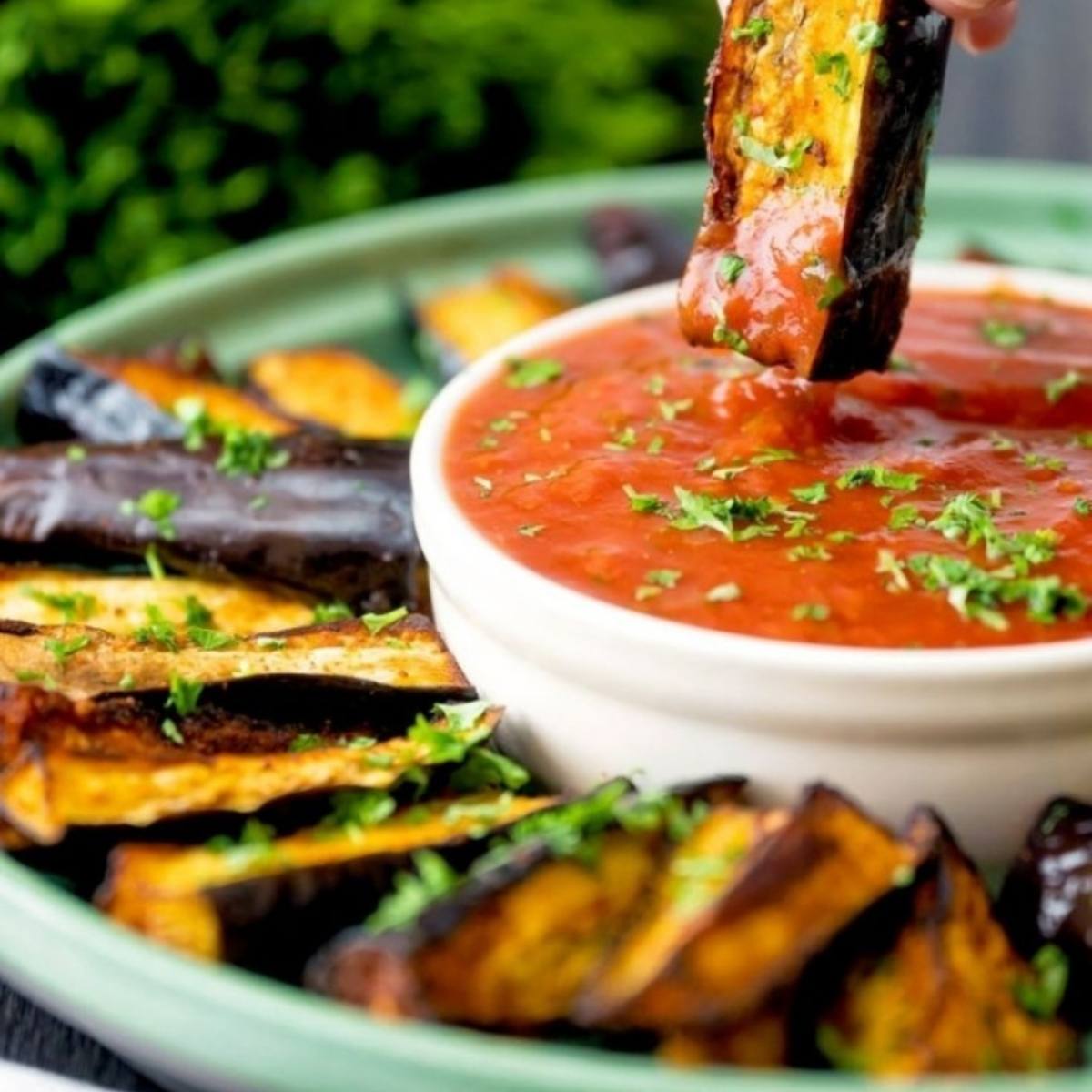 A hand dipping a crispy, golden-brown eggplant fry into a bowl of marinara sauce. The eggplant fries look delicious and perfect for dipping.