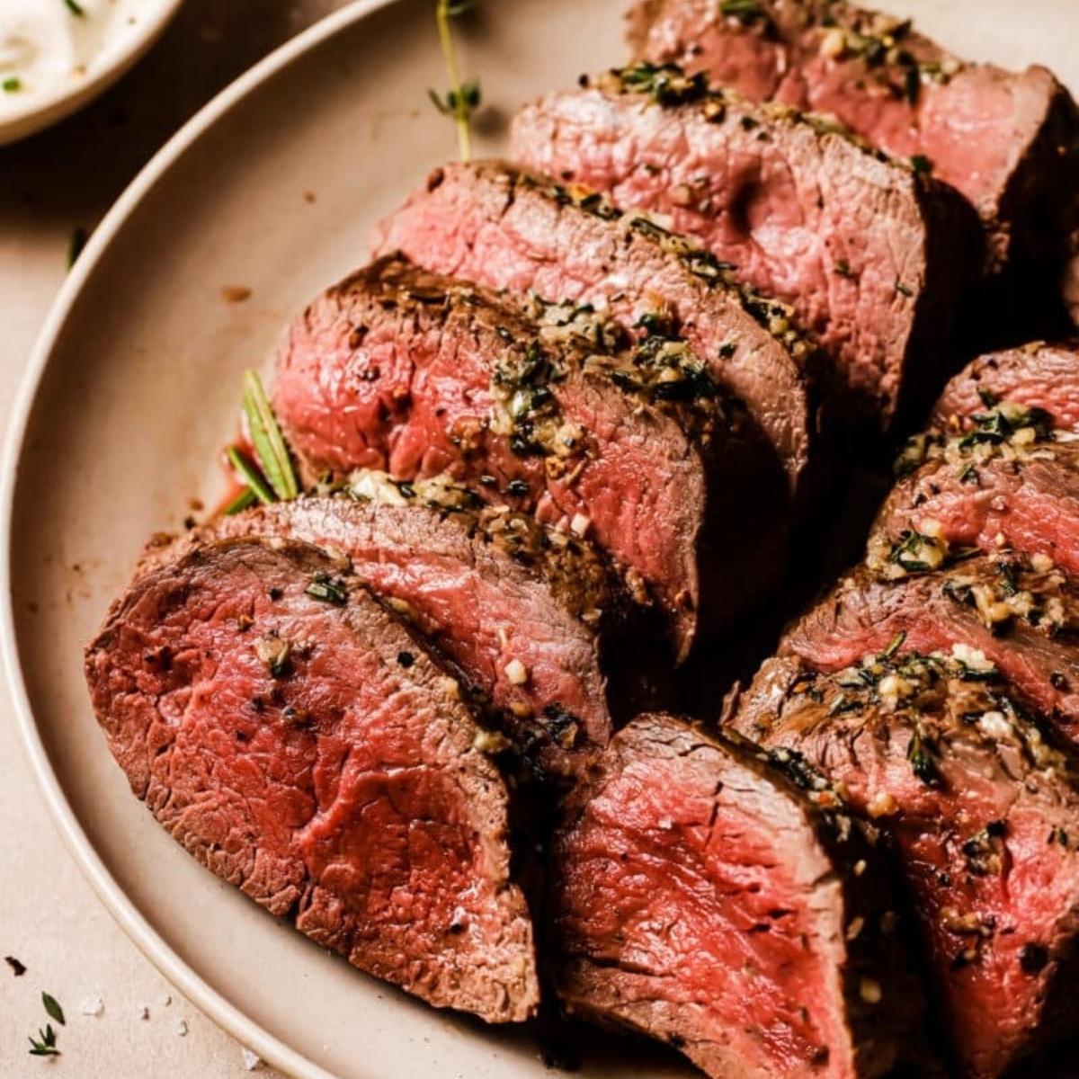 A close-up of a plate of perfectly cooked and sliced beef tenderloin. The meat is medium-rare, juicy, and topped with a crust of herbs and spices.







