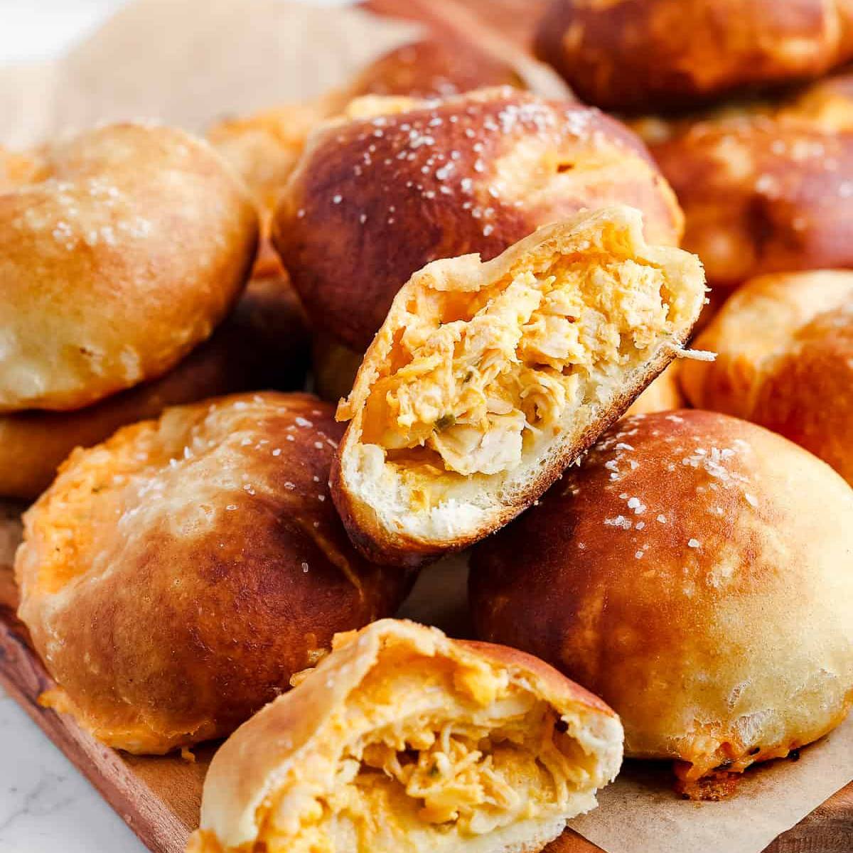 A close-up of several golden-brown pretzel buns filled with a creamy buffalo chicken filling. One of the buns is cut in half, revealing the flavorful filling inside.








