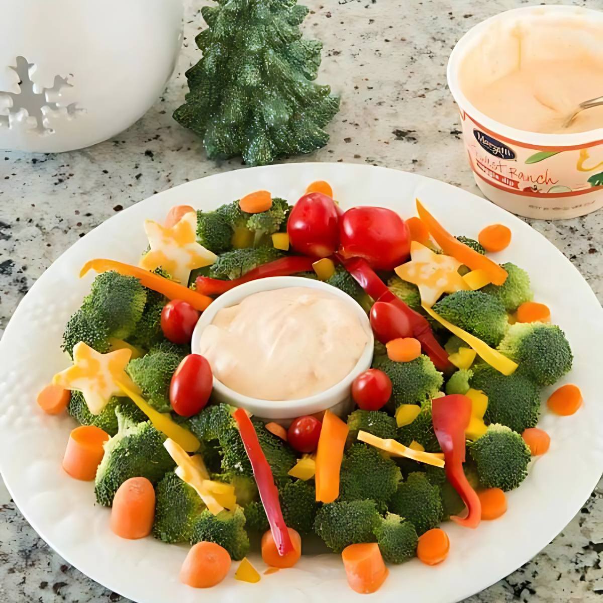 A festive vegetable platter arranged in the shape of a Christmas wreath. The wreath is made up of broccoli florets, cherry tomatoes, carrot sticks, and cheese stars. A small bowl of ranch dressing is placed in the center.