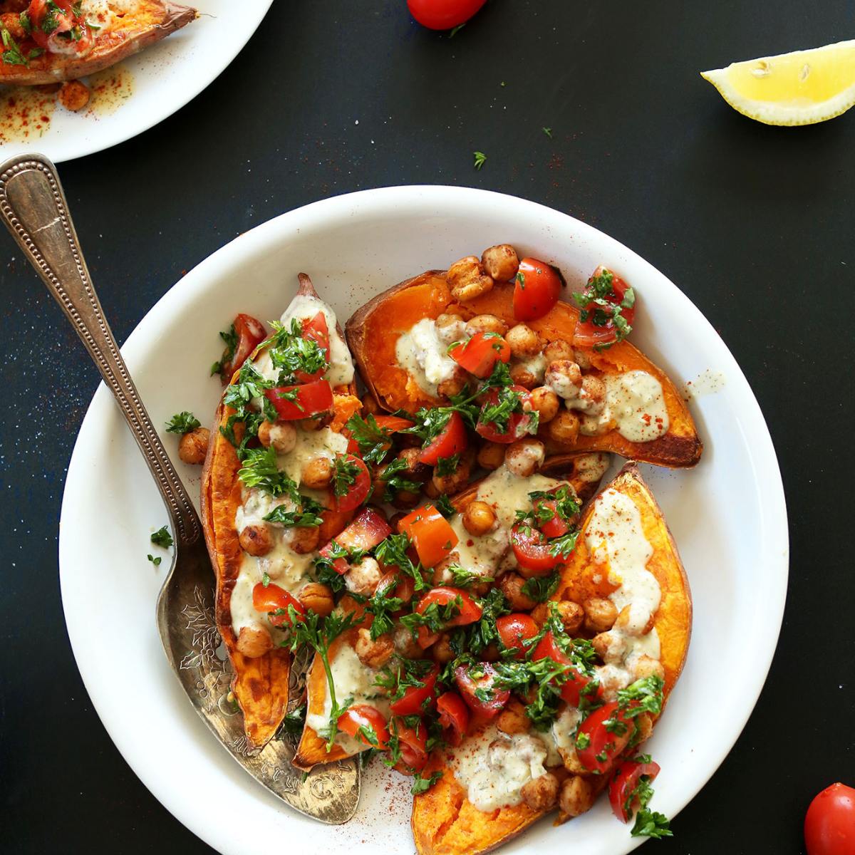 A plate of roasted sweet potato wedges topped with chickpeas, tomatoes, tahini sauce, and fresh herbs.