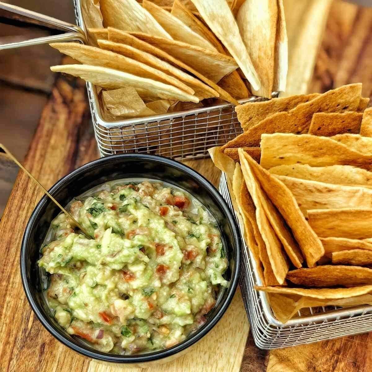 A bowl of guacamole with a spoon and a basket of tortilla chips on a wooden table. This looks like a perfect snack or appetizer for a party!