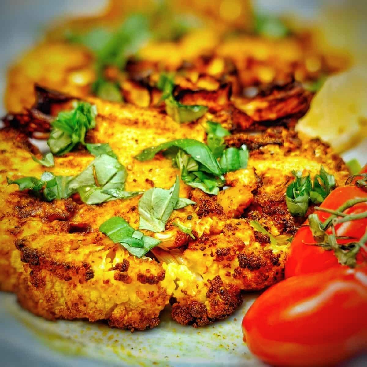 A close-up of crispy, golden-brown cauliflower steaks. The cauliflower is seasoned and cooked to perfection, and it's garnished with fresh basil leaves and cherry tomatoes.