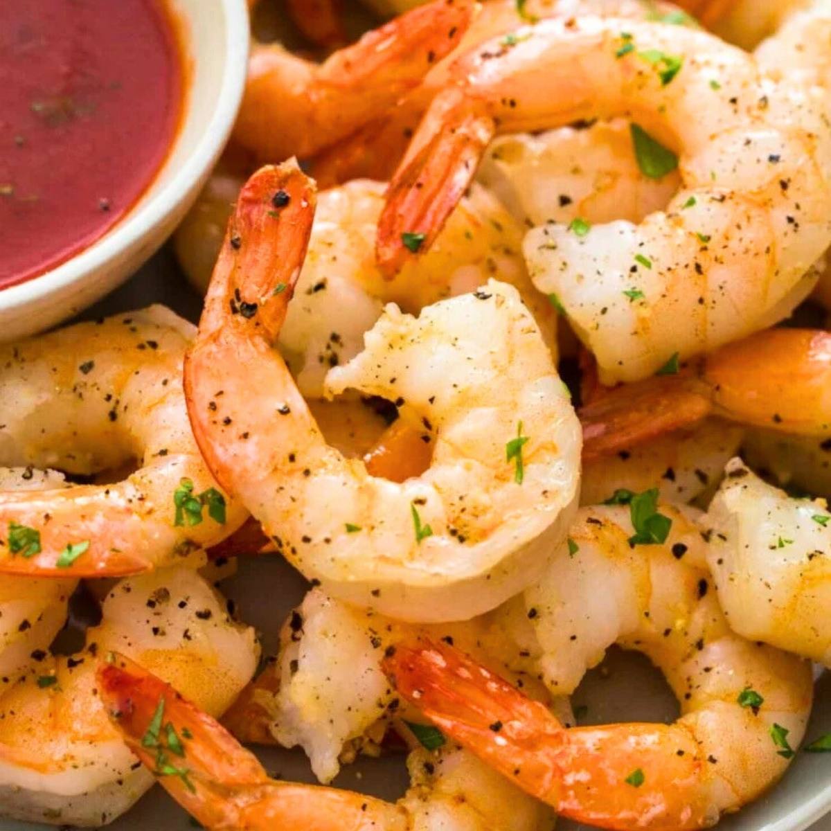 A plate of cooked shrimp with a bowl of cocktail sauce. The shrimp are plump and juicy-looking, and they appear to be seasoned with salt and pepper.