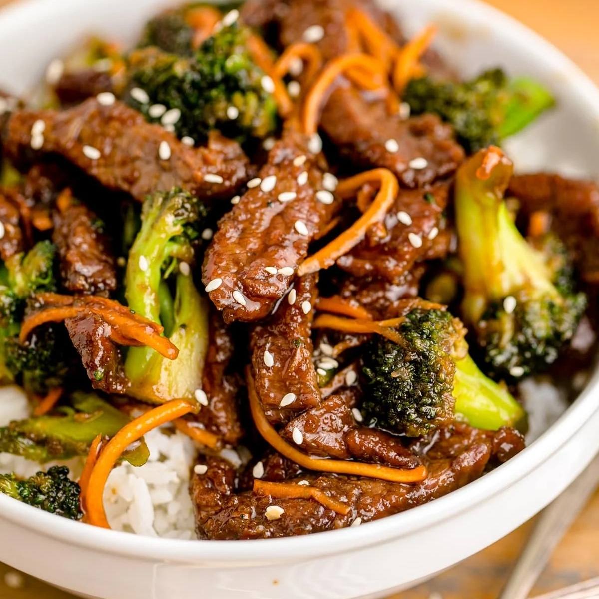 A bowl of Mongolian beef and broccoli cooked in an air fryer. The beef is tender and coated in a rich, savory sauce, and it's served with broccoli florets, shredded carrots, and a bed of white rice. Sesame seeds are sprinkled on top