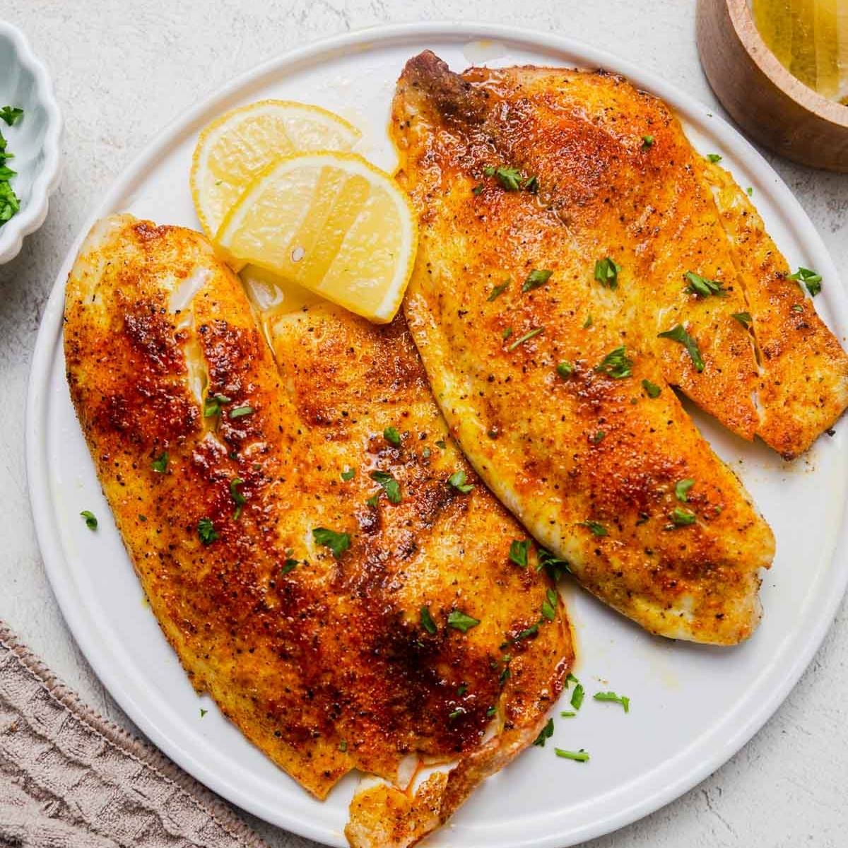 A plate of three perfectly cooked tilapia fillets. The fillets are golden brown and seasoned with herbs and spices. They are served with lemon wedges and fresh parsley.