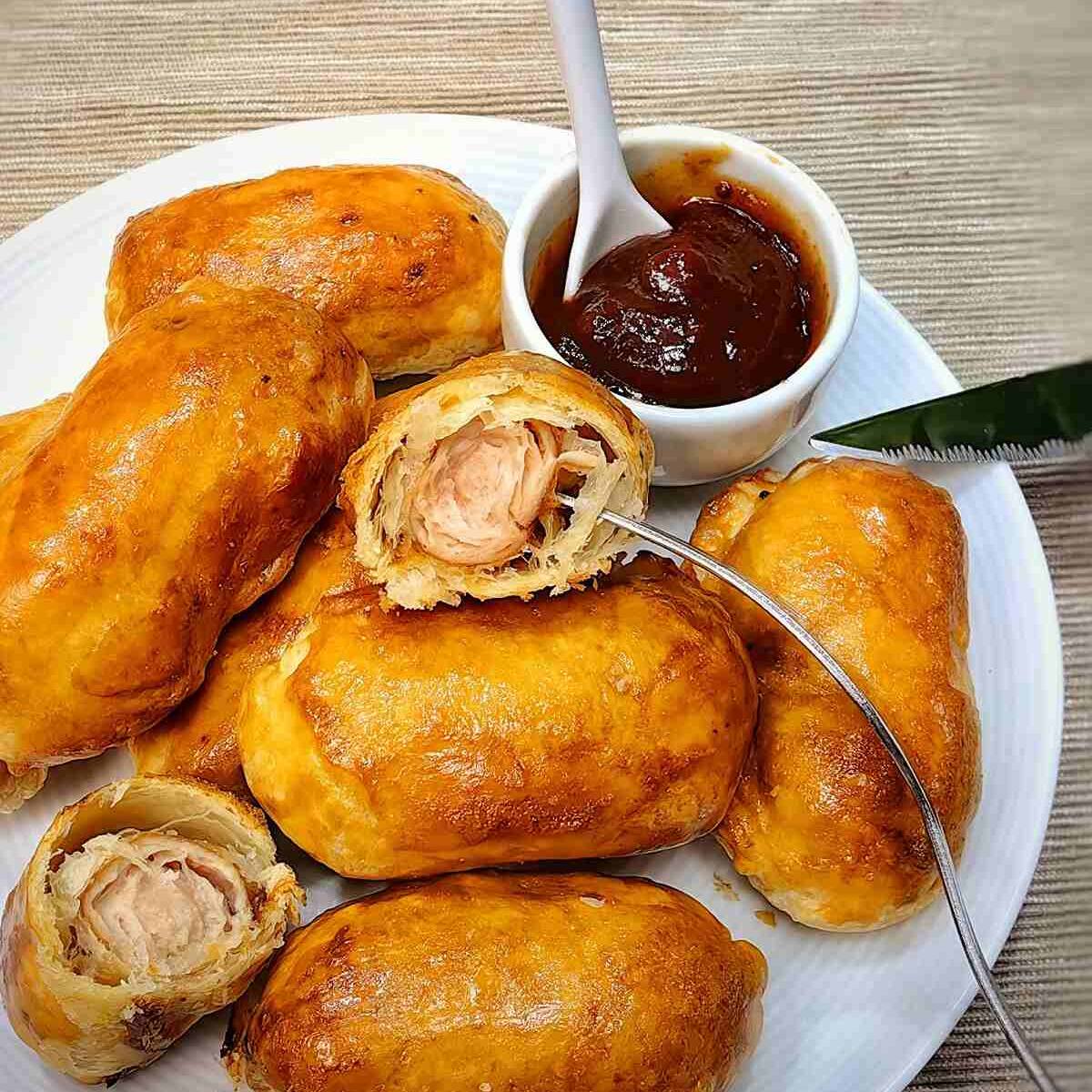 A plate of golden-brown sausage rolls with a side of ketchup. One of the rolls has been cut open to reveal the juicy sausage inside.