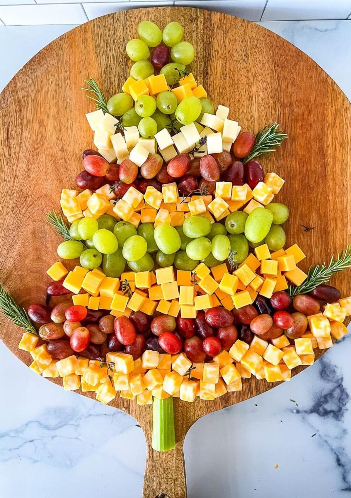 A festive Christmas tree-shaped charcuterie board filled with cheese cubes and grapes. The board is decorated with rosemary sprigs.







