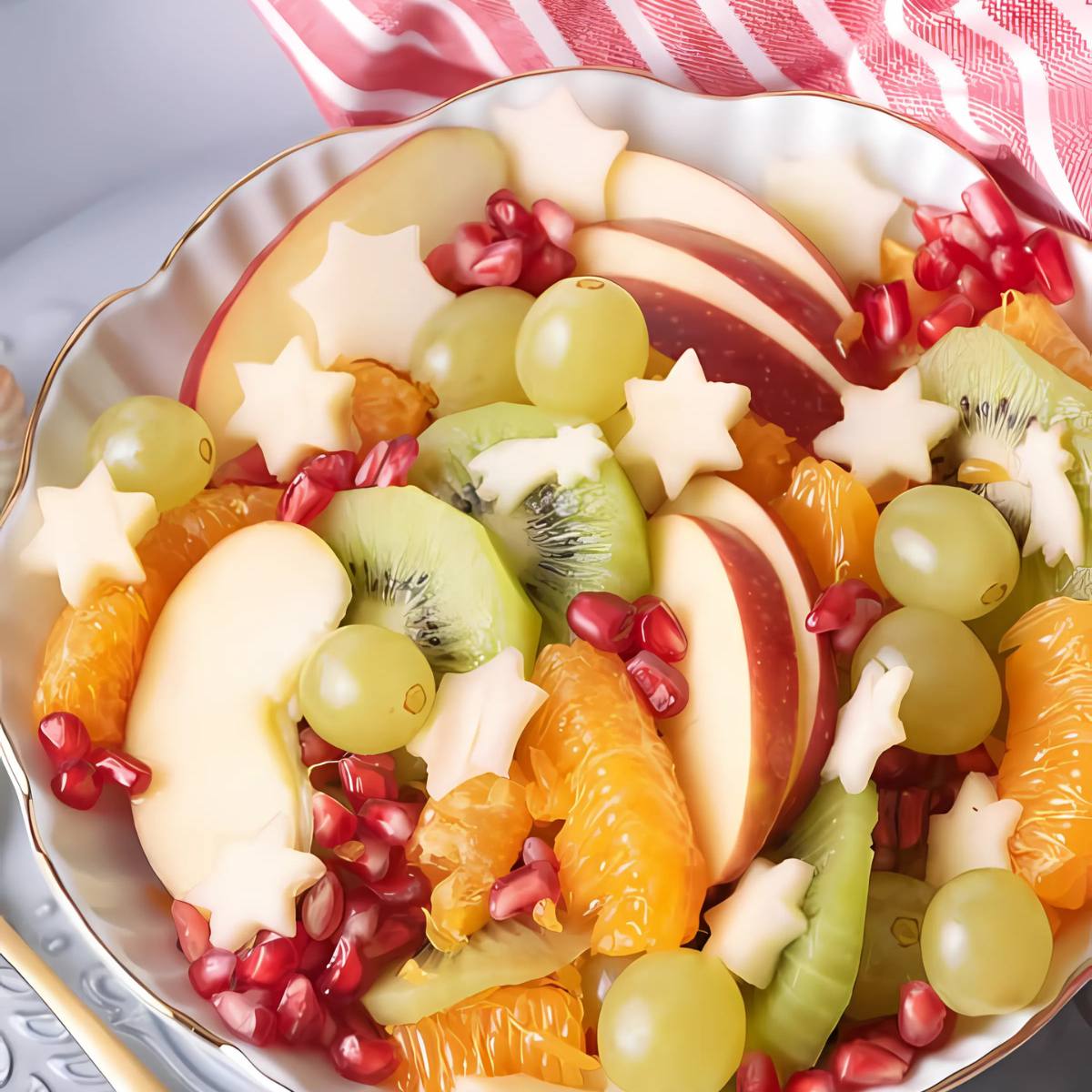 A bowl of colorful fruit salad. The salad includes apples, kiwi, grapes, mandarin oranges, and pomegranate seeds. Some of the apple slices are cut into star shapes.