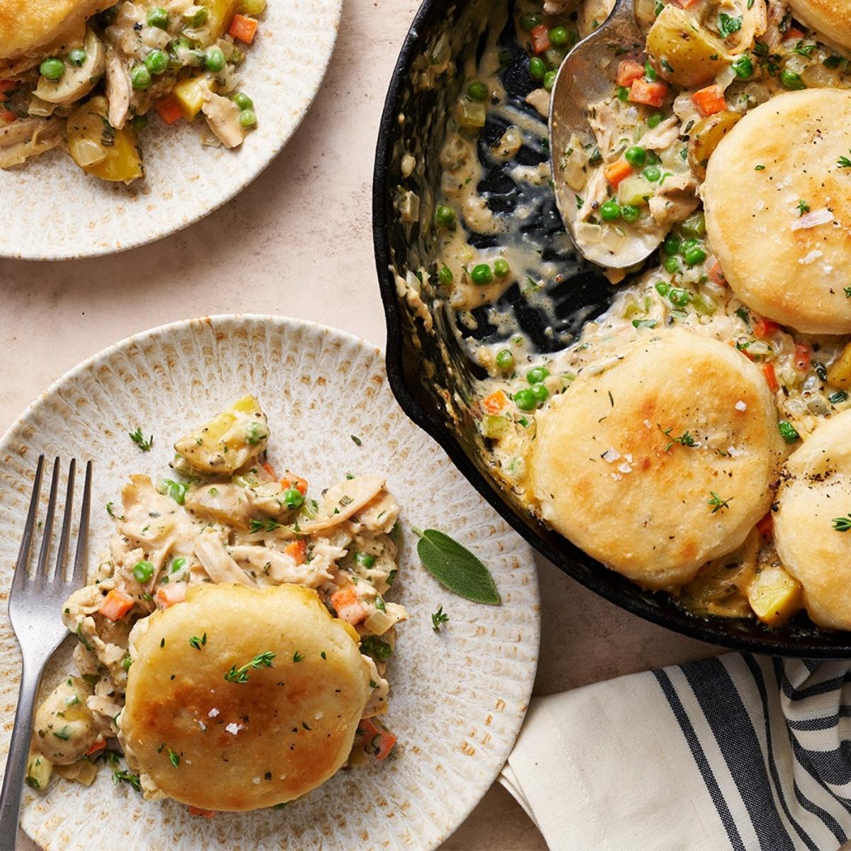 A top-down view of a cast iron skillet filled with a creamy chicken pot pie. The pot pie is topped with fluffy biscuits and served with additional biscuits on the side.