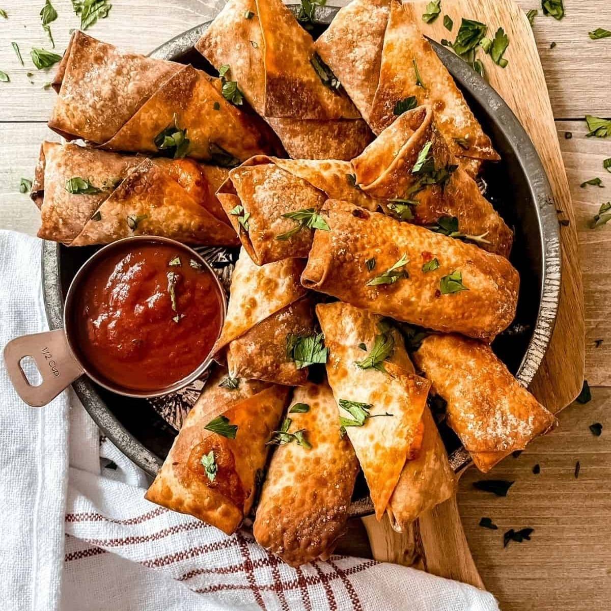 A platter of crispy, golden-brown egg rolls arranged neatly. They are sprinkled with fresh parsley and served with a small bowl of marinara sauce for dipping.