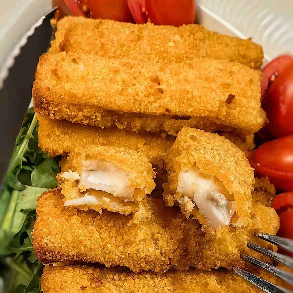 A close-up of crispy, golden-brown fish fingers on a plate. A fork is lifting one of the fish fingers, showing the flaky white fish inside. There are also some cherry tomatoes and arugula on the plate.