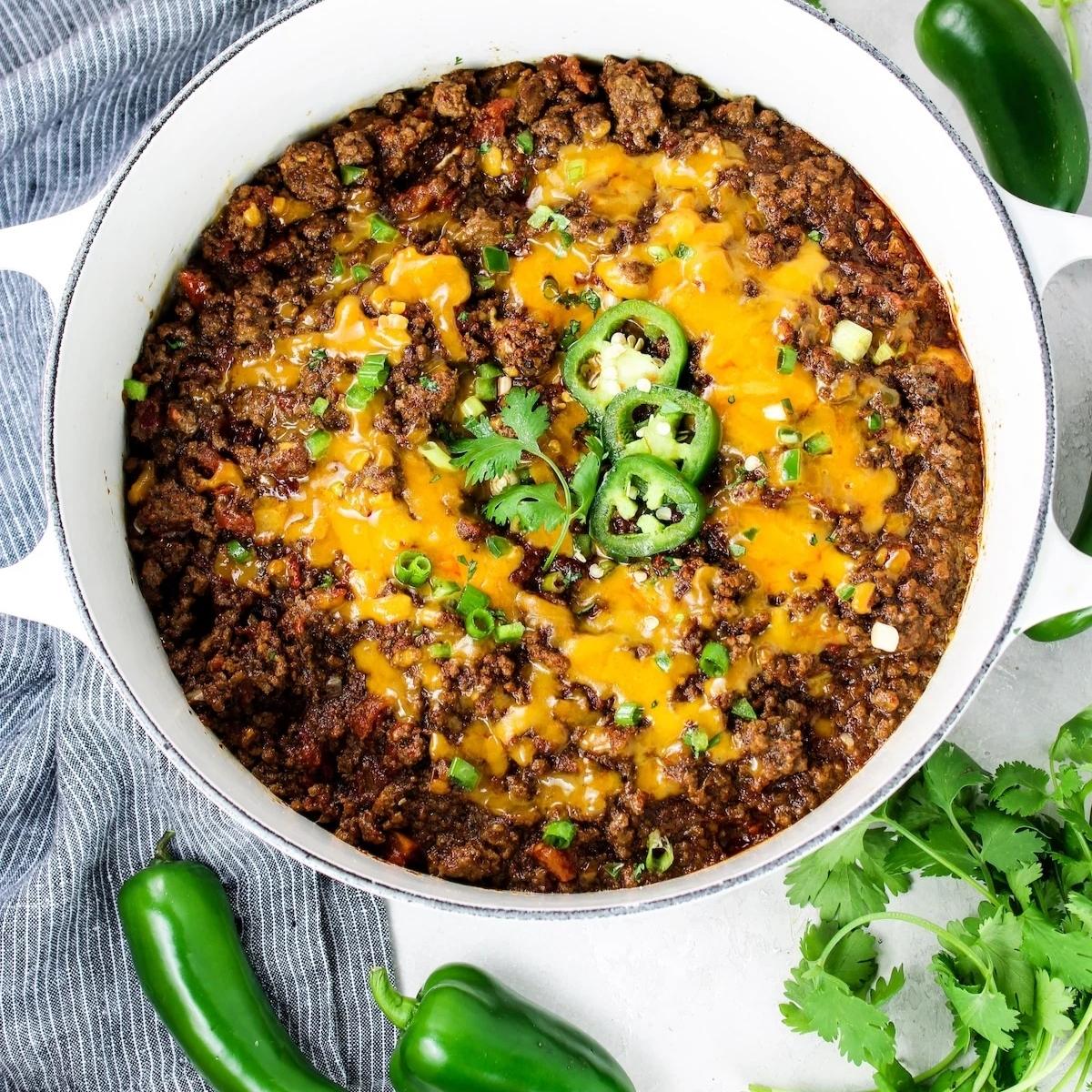 A hearty bowl of chili topped with melted cheddar cheese, fresh cilantro, and sliced jalapeños. The chili appears to have a rich, meaty texture and is ready to be served.