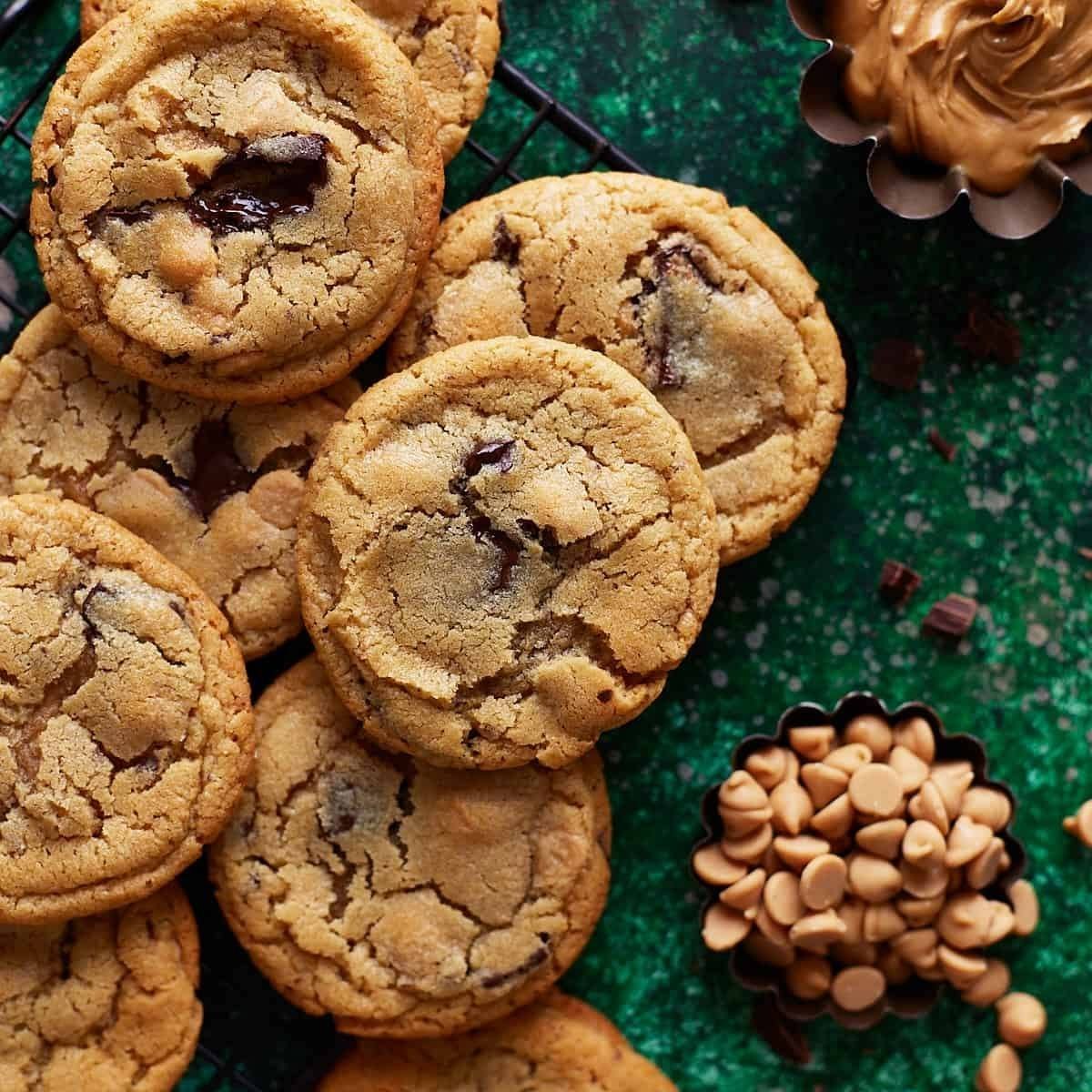 A close-up of a stack of golden-brown chocolate chip cookies. The cookies are soft and chewy-looking, with visible chocolate chips. There are also bowls of butterscotch chips and peanut butter in the background.