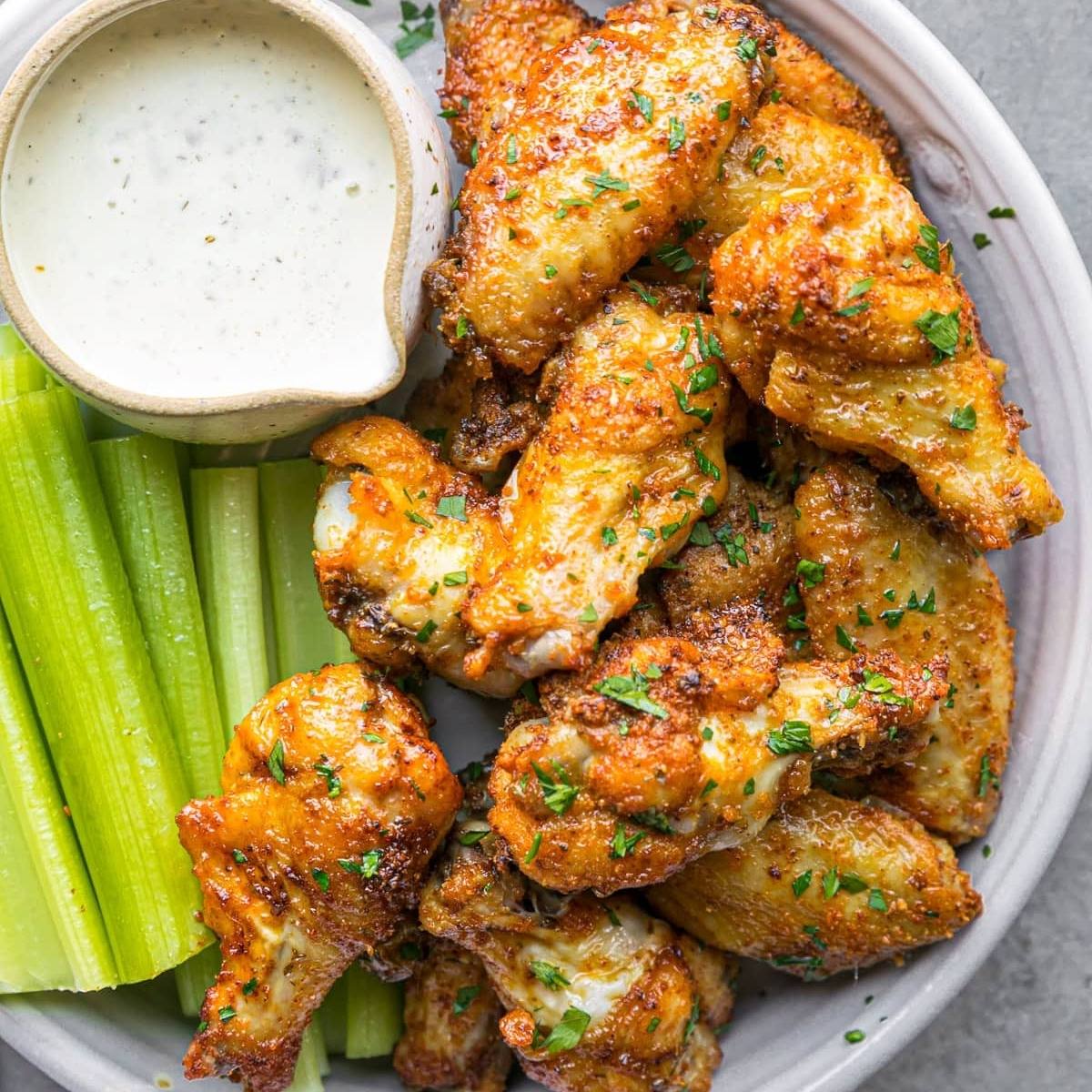 A plate of crispy, golden-brown chicken wings arranged neatly. They are sprinkled with fresh parsley and served with a side of celery sticks and a bowl of ranch dressing for dipping.