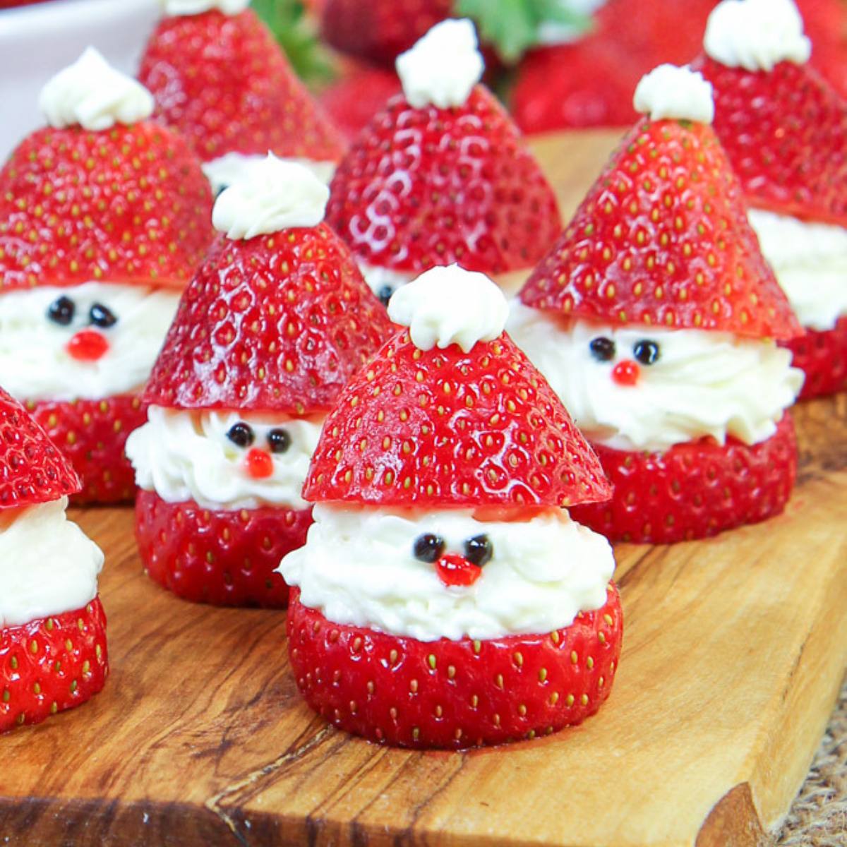 A wooden board filled with adorable Santa Claus strawberries. Each strawberry is topped with whipped cream to form a hat, with chocolate chips and red sprinkles used to create the face.