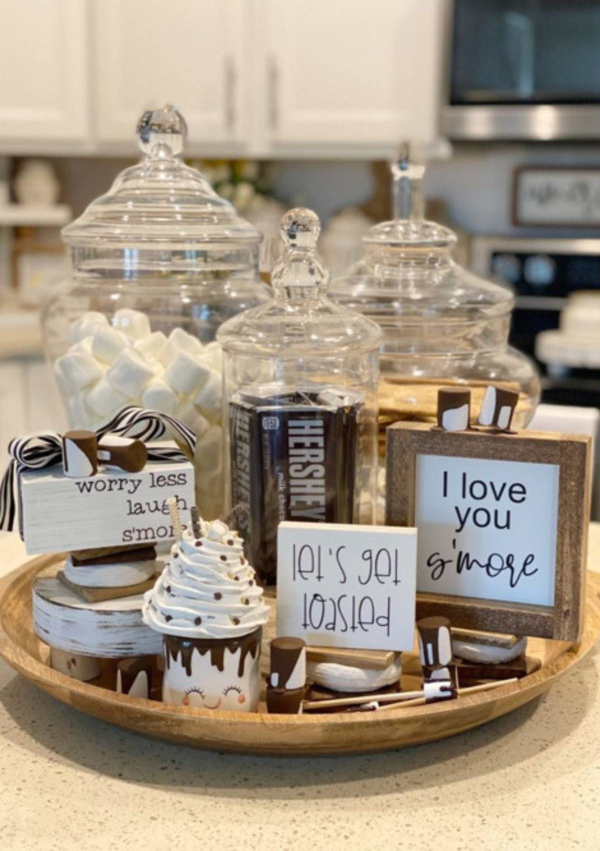 A festive s'mores station with glass jars filled with marshmallows, chocolate bars, and graham crackers. There are also wooden signs with s'mores-themed messages and a cute s'mores-themed mug.