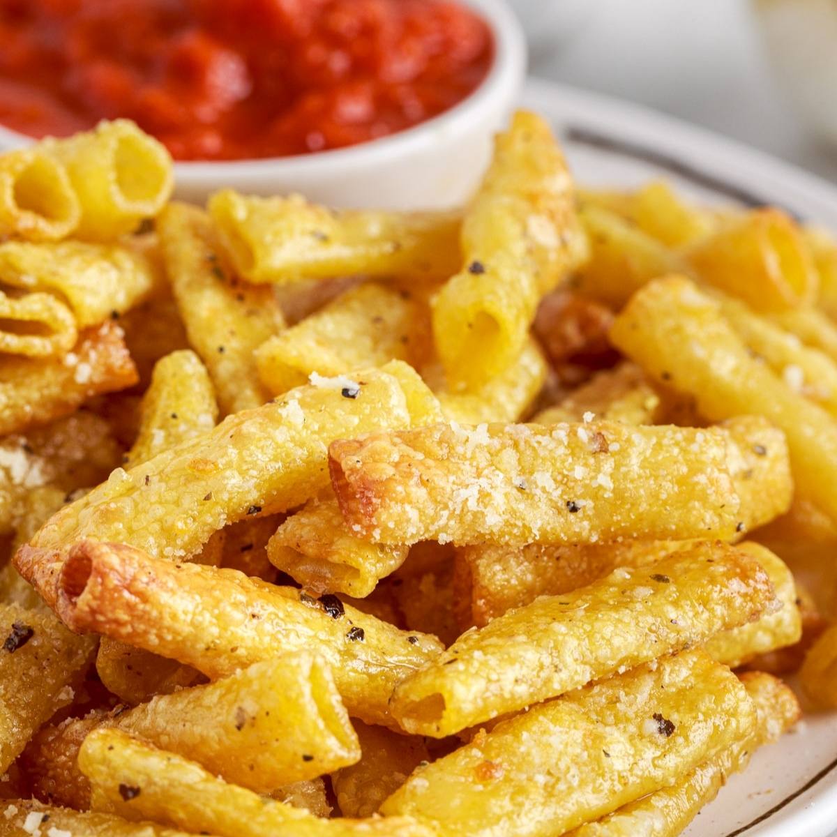 A plate of crispy, air fried pasta chips. The pasta chips are golden brown and seasoned with herbs and spices. There is a bowl of marinara sauce in the background, ready for dipping.