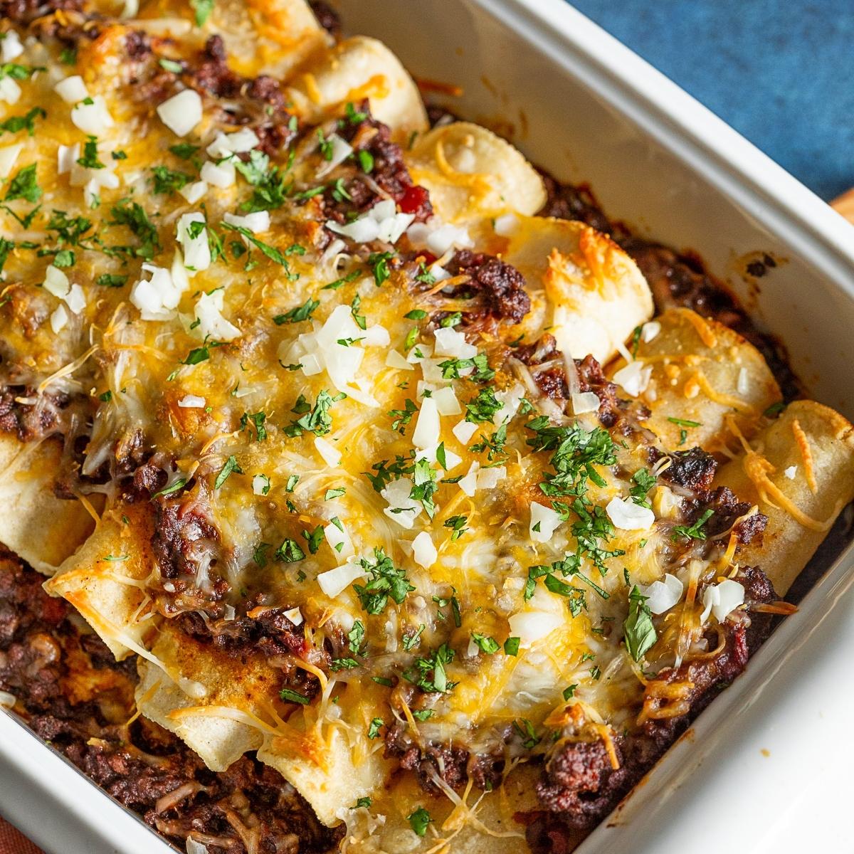 A close-up of a baking dish filled with golden-brown enchiladas. The enchiladas are generously topped with melted cheese, chopped onions, and fresh parsley.