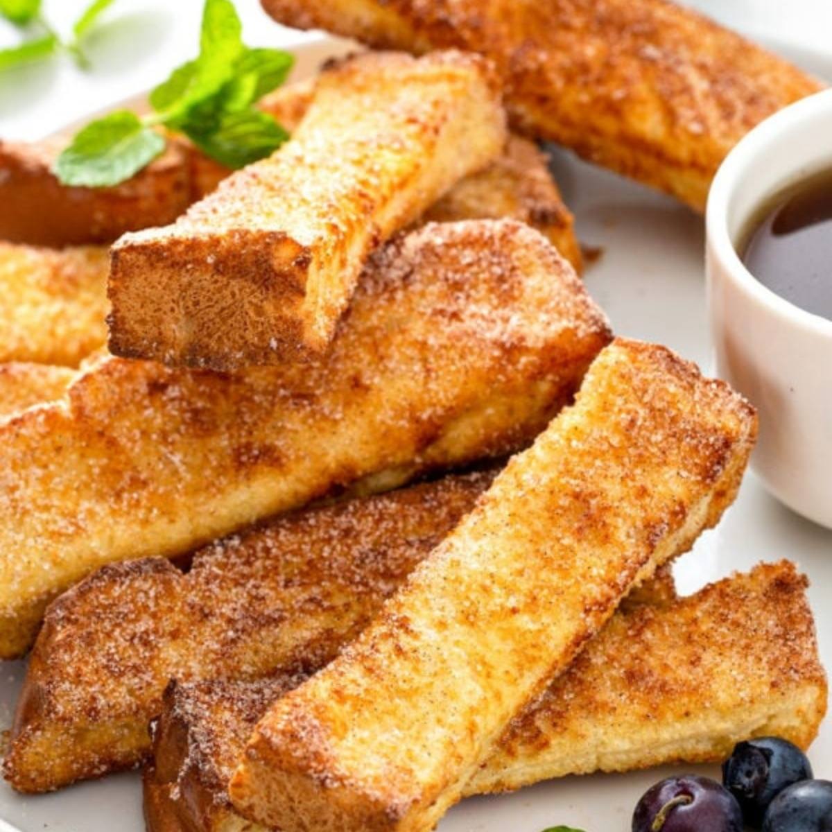 A plate of golden-brown French toast sticks. The sticks are coated in cinnamon sugar and look delicious. There is a bowl of syrup and some fresh blueberries on the side.