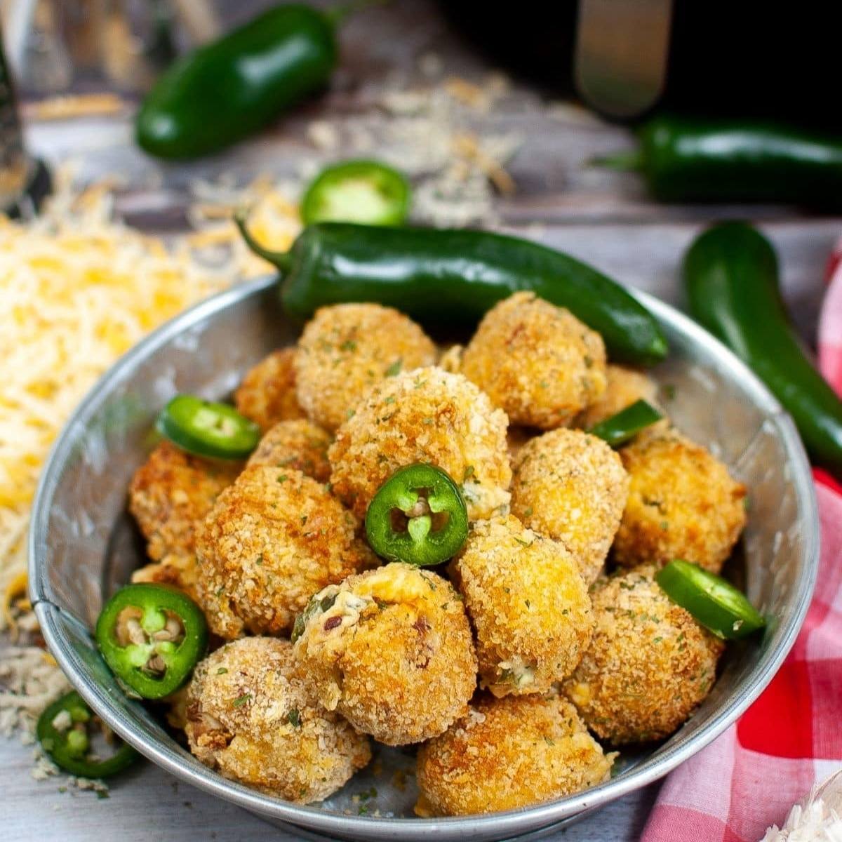 A bowl of golden-brown, breaded jalapeno poppers. The poppers are filled with cheese and look crispy and delicious. There are fresh jalapenos and shredded cheese in the background.