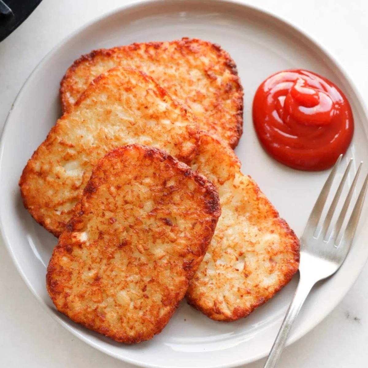 A plate of golden-brown, crispy hash browns with a dollop of ketchup and a fork. The hash browns look perfectly cooked and ready to be enjoyed for breakfast or brunch.