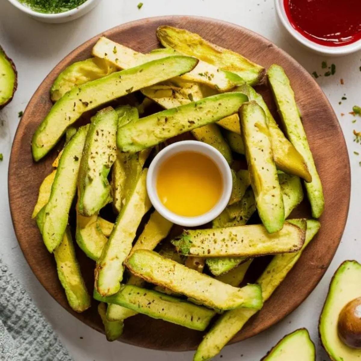 A plate of crispy, baked avocado fries. The fries are golden brown and seasoned with herbs. There are bowls of dipping sauces, including ketchup and a creamy sauce, on the side.