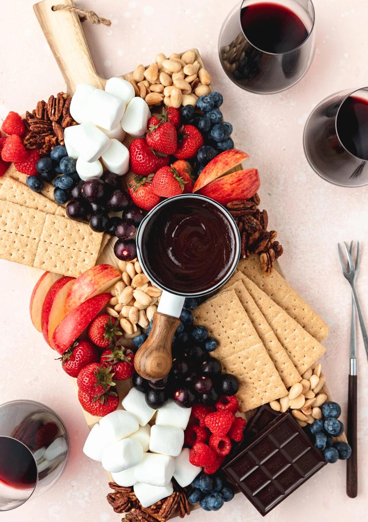 A festive chocolate fondue board filled with an assortment of fruits, nuts, and marshmallows, ready to be dipped into the melted chocolate. There are also glasses of red wine and a chocolate bar on the board.