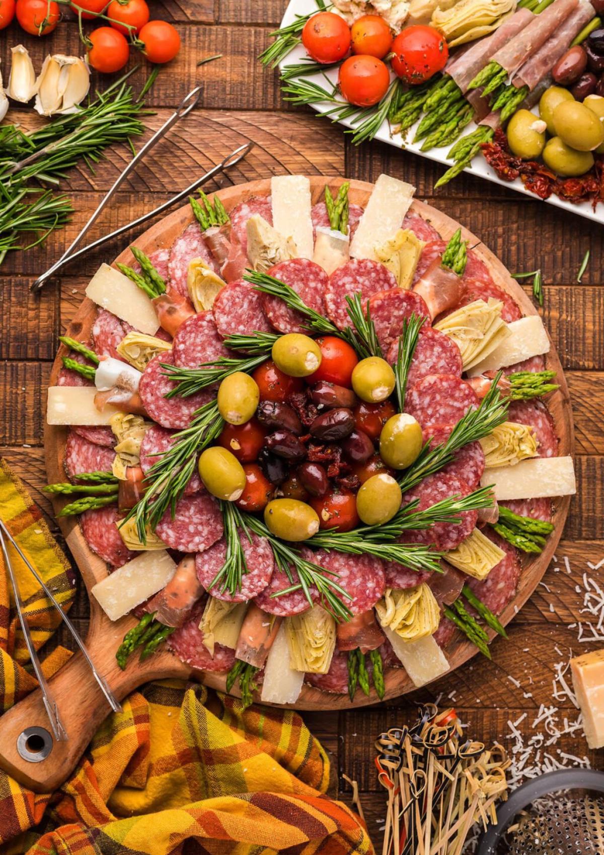 A festive charcuterie board featuring a variety of meats, cheeses, olives, artichoke hearts, asparagus, and tomatoes. The board is decorated with fresh rosemary sprigs.