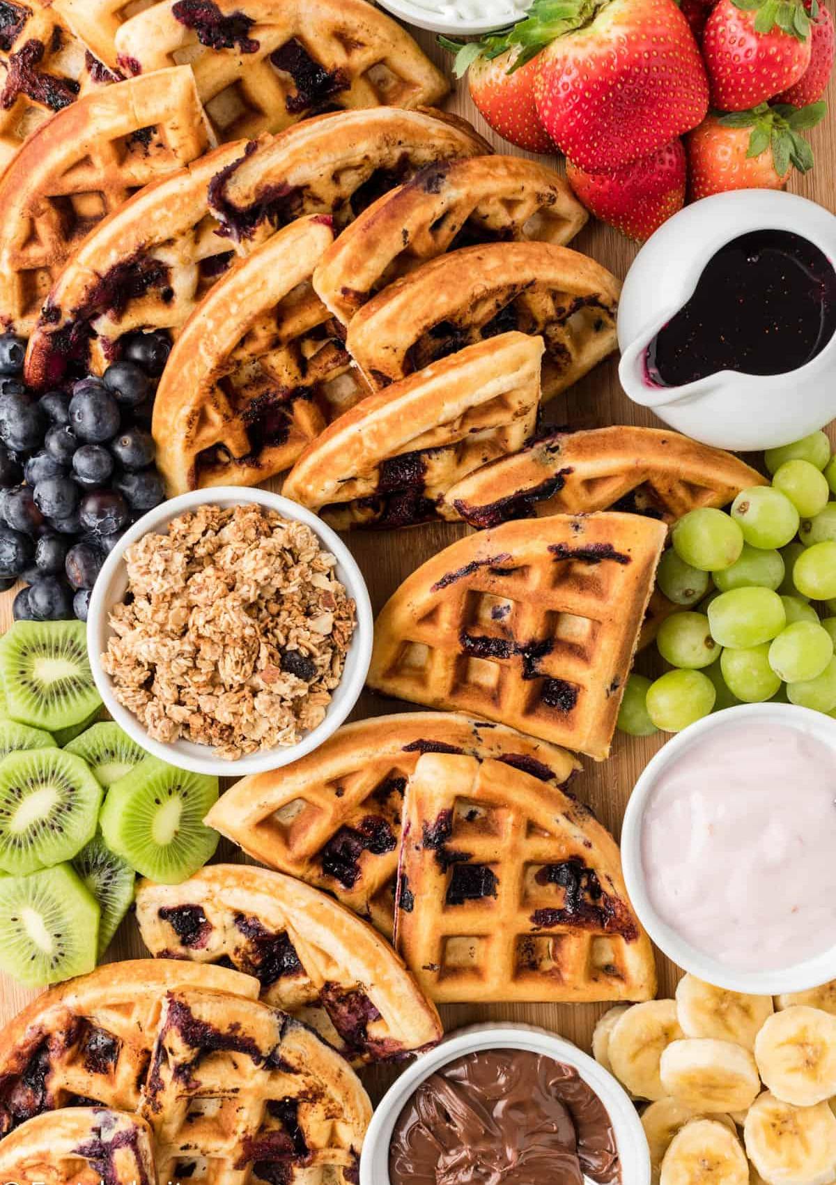 The board includes waffles, fresh fruit like strawberries, blueberries, grapes, kiwi, and bananas, as well as yogurt, granola, chocolate hazelnut spread, and syrup.