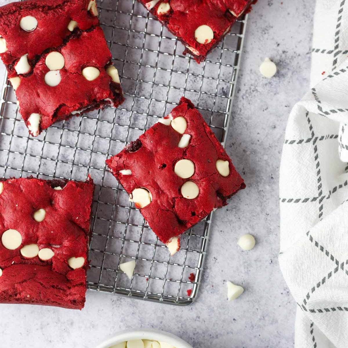 A cooling rack with squares of red velvet brownies topped with white chocolate chips.