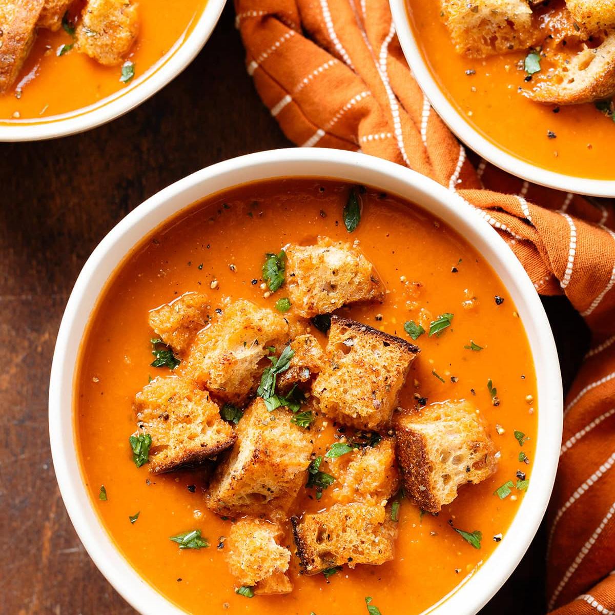 A bowl of creamy tomato soup topped with croutons and fresh parsley. The soup is a vibrant orange color and the bowl is white. The soup is resting on an orange and white striped napkin.