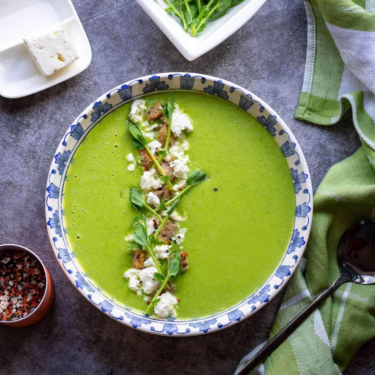 A bowl of vibrant green pea soup garnished with feta cheese, croutons, and pea shoots. The bowl has a decorative blue and white pattern.