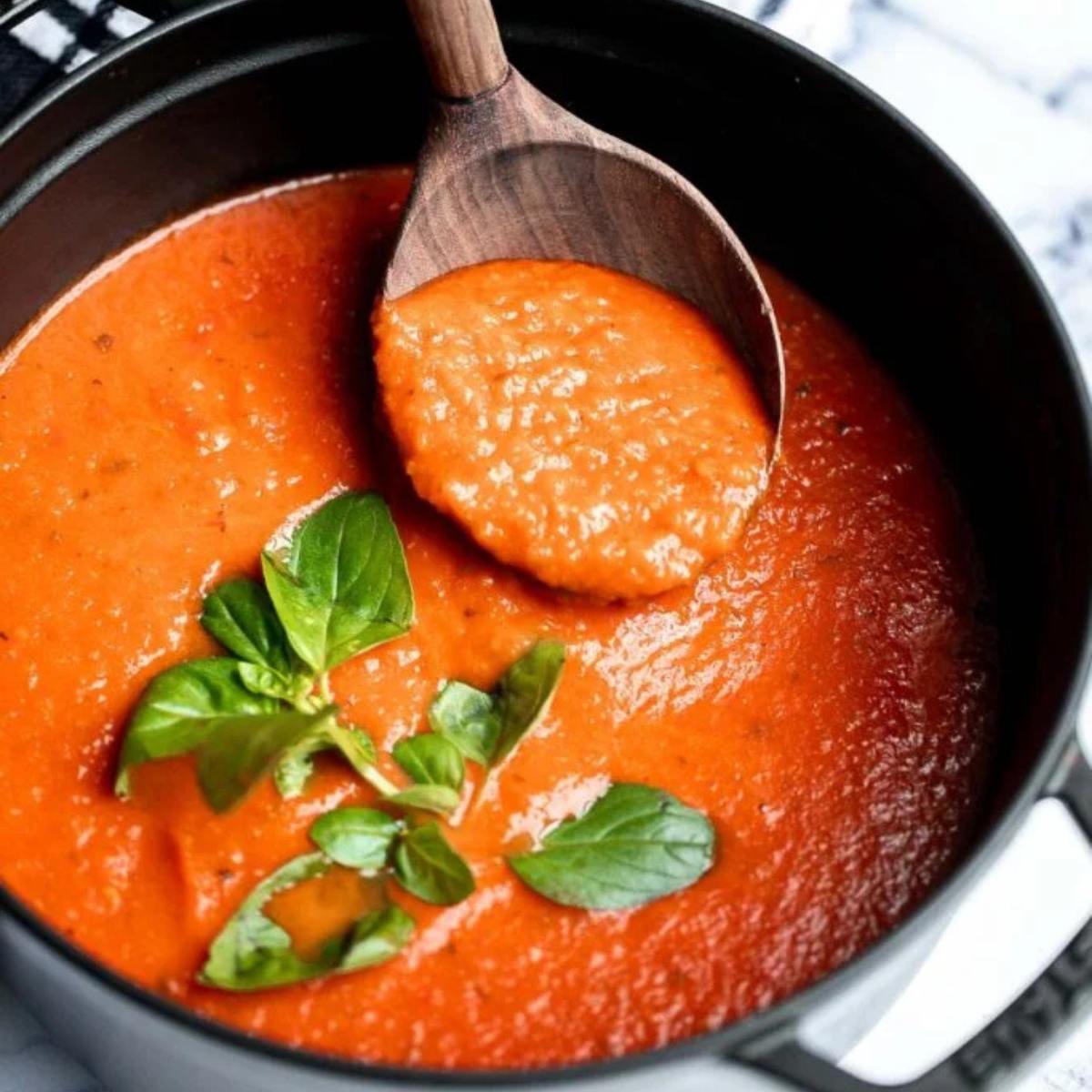 A pot of vibrant red tomato soup with a wooden spoon scooping some out. Fresh basil leaves are sprinkled on top.